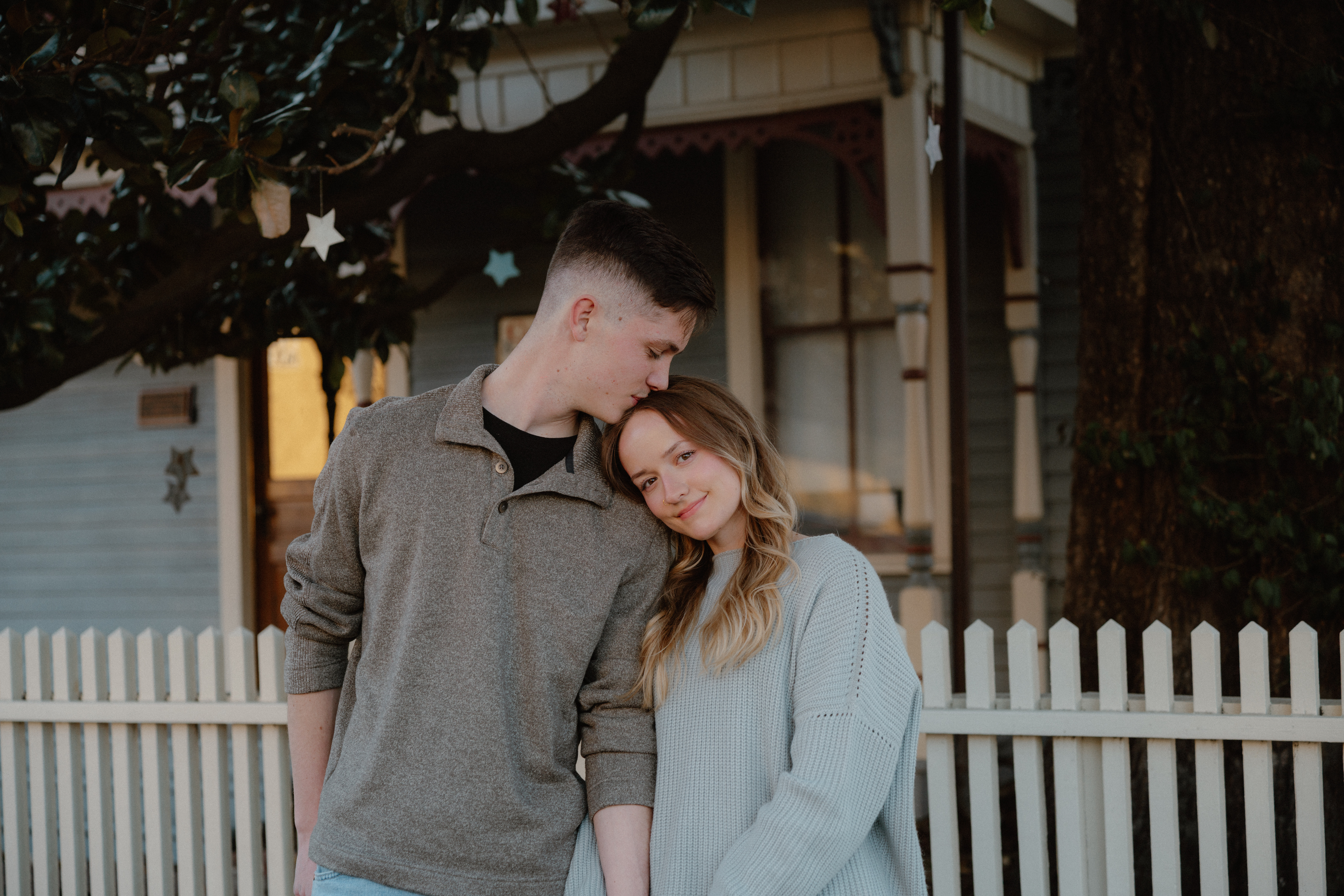 Couple standing in front of fence with the man kissing the woman's head
