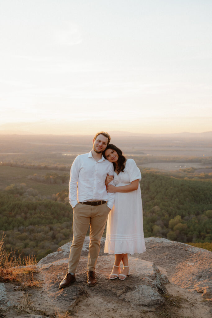 Couple holding each other on a mountain while looking at the camera in front of the sunset 